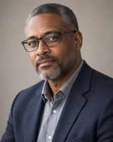 A man in his fifties with close-cropped greying hair and dark-framed glasses, in a navy jacket, photographed from the chest up against a plain warm grey background.