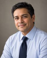 A man in his thirties with short dark hair, in a light blue shirt and dark tie, photographed from the chest up against a plain warm grey background.