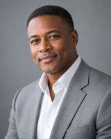 A man in his forties with closely cropped hair, in a light grey jacket over an open-collar shirt, photographed from the chest up against a plain cool grey background.