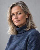 A woman in her fifties with shoulder-length ash blonde hair, in a slate blue roll-neck jumper, photographed from the chest up against a plain pale grey background.