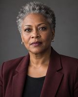 A woman in her fifties with short grey hair, in a dark burgundy blazer, photographed from the chest up against a plain dark grey background.