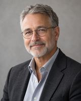 A man in his sixties with a grey beard and wire-rimmed glasses, in a charcoal jacket, photographed from the chest up against a plain pale background.