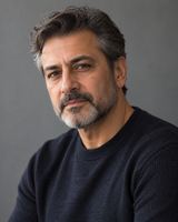 A man in his fifties with greying hair and a short beard, in a dark crew-neck jumper, photographed from the chest up against a plain slate grey background.